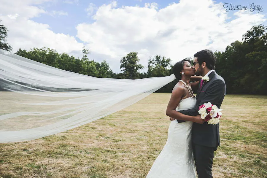 Les mariés s’embrassent dans le parc, le voile de la mariée déployé par le vent sur toute la largeur de l’image
