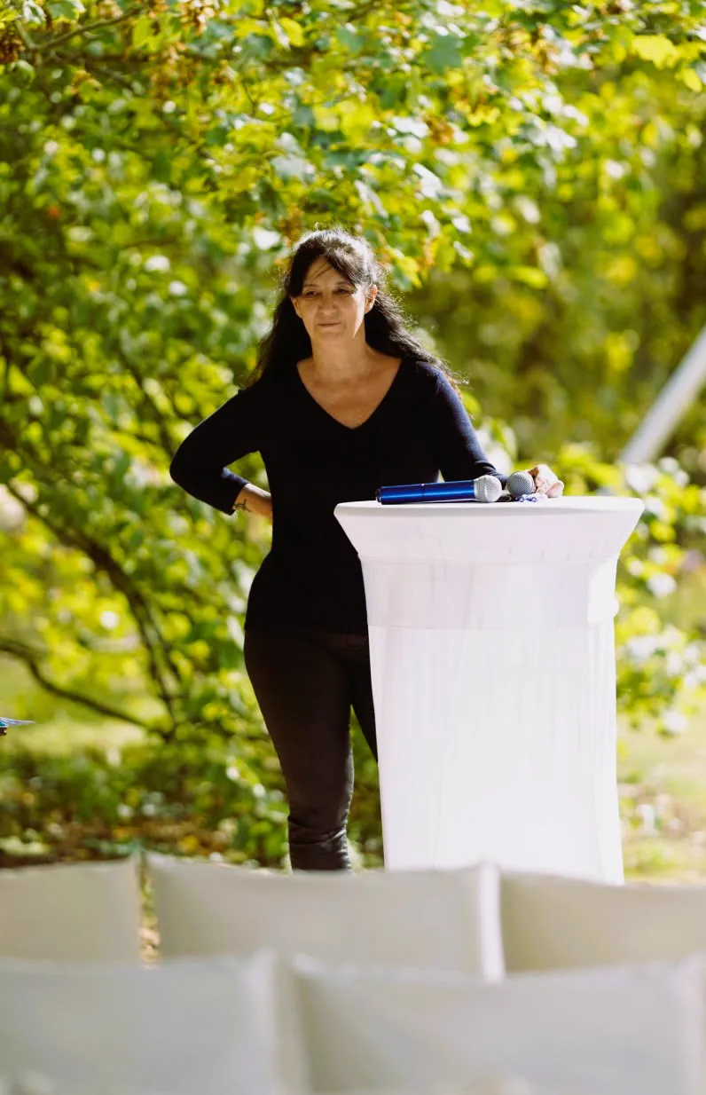 Anne Biotteau debout au pupitre pendant une c&eacute;r&eacute;monie en plein air, les micros pos&eacute;s devant elle