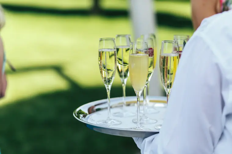 Un serveur ganté de blanc porte un plateau argenté de flûtes de champagne, en extérieur sur une pelouse