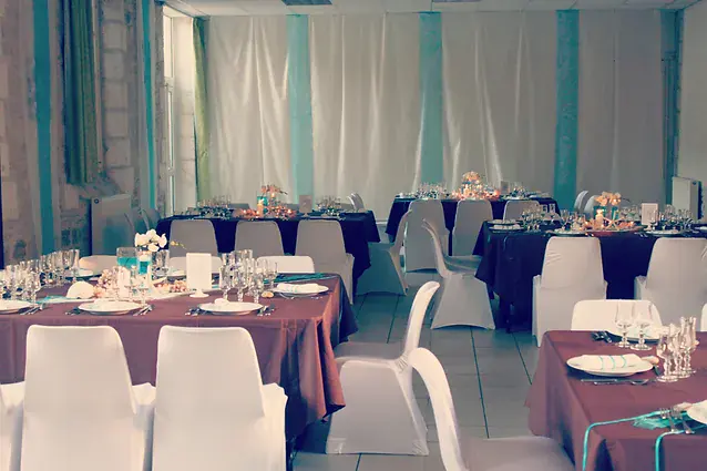 La salle habill&eacute;e de voilages turquoise et blancs, longues tables en chocolat et housses blanches