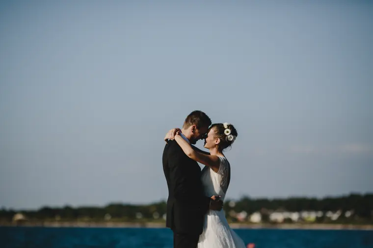 Un couple front contre front au bord de l’eau, seuls, la mariée coiffée de fleurs blanches