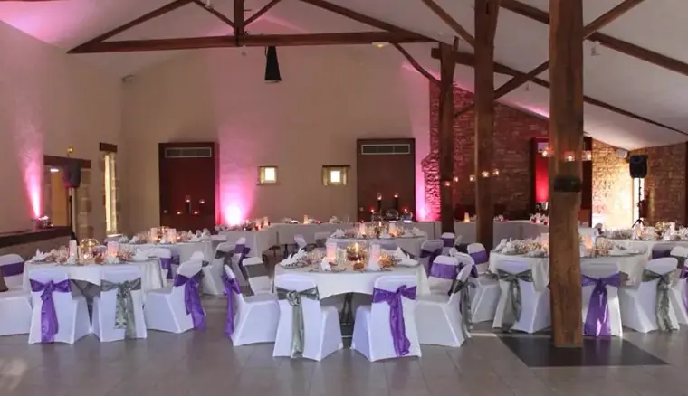 La salle sous la charpente : pierres apparentes, poteaux de bois, tables rondes et murs lavés de rose
