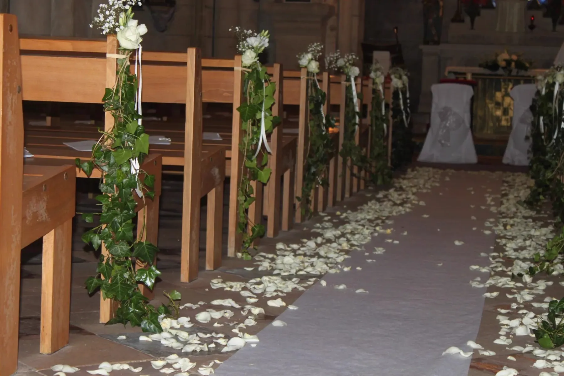 L&rsquo;all&eacute;e centrale d&rsquo;une &eacute;glise, guirlandes de lierre et roses blanches aux bancs, p&eacute;tales blancs au sol