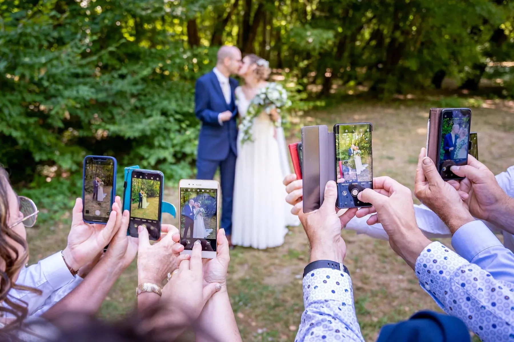 Les mariés s’embrassent dans un parc en plein été, le marié en costume bleu marine, photographiés par leurs invités