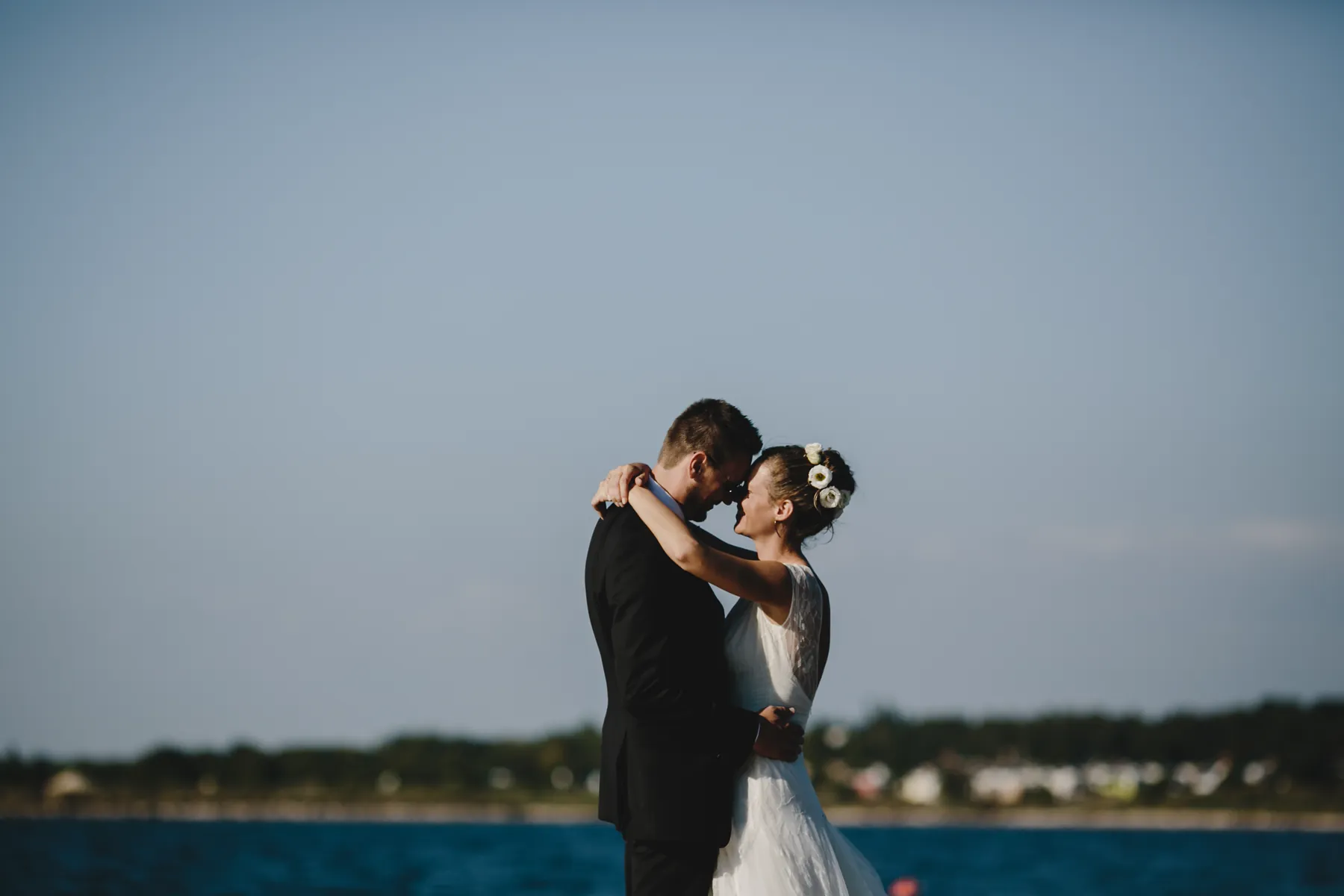 Un couple front contre front au bord de l’eau, seuls, la mariée coiffée de fleurs blanches