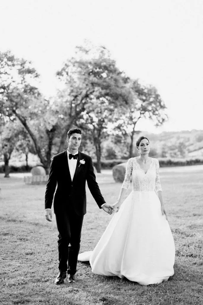 Les mariés marchent main dans la main dans un parc, en noir et blanc, le marié en smoking avec une seule boutonnière blanche au revers