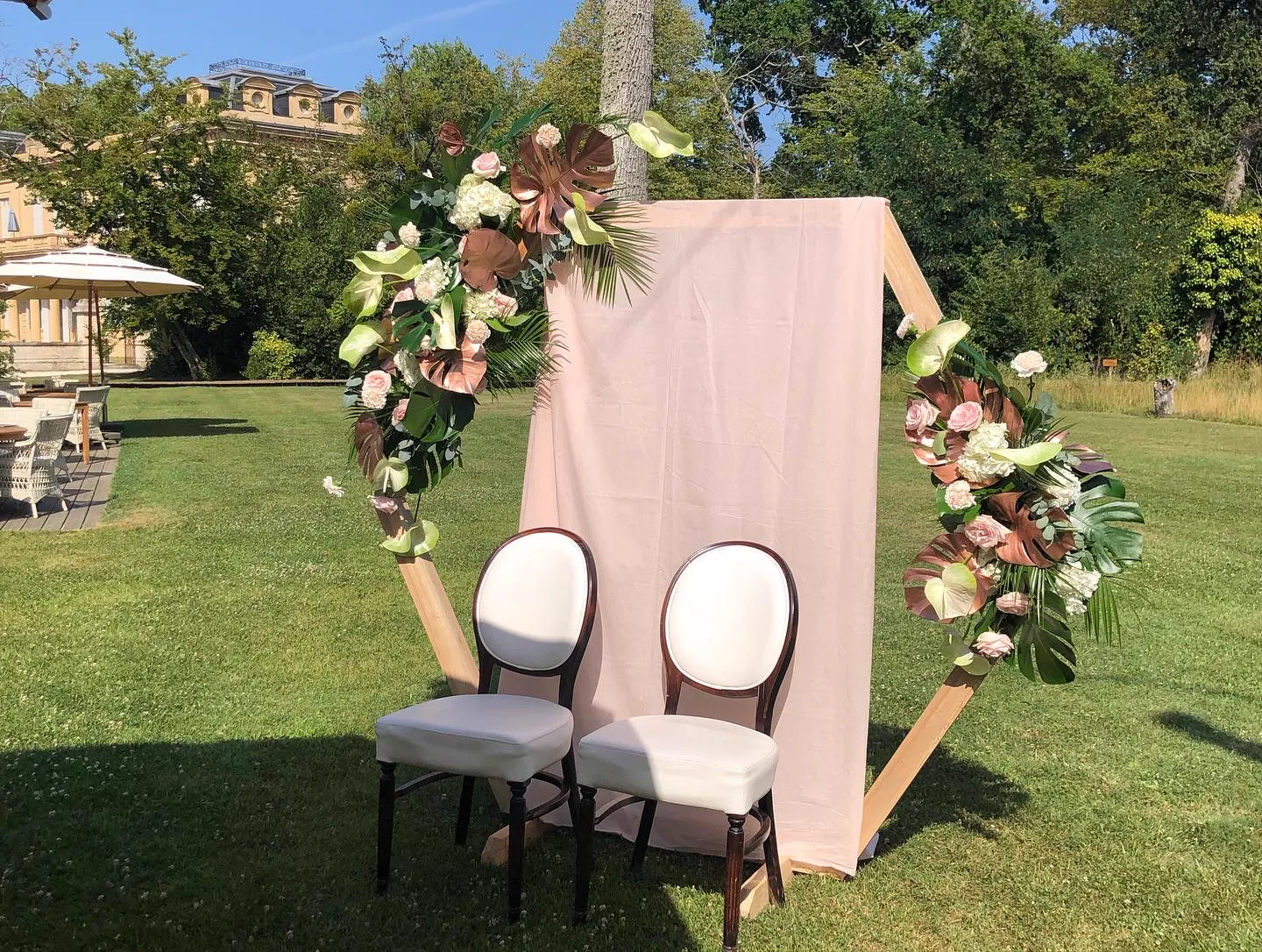 Une arche hexagonale de bois clair, drapée de rose poudré et fleurie d’hortensias et de feuillages cuivrés, sur la pelouse devant le château
