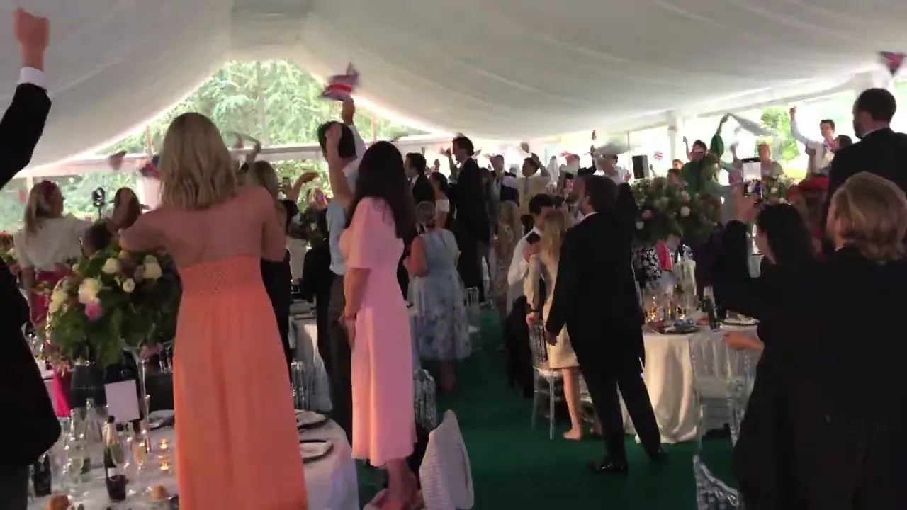 Les invit&eacute;s debout agitent de petits drapeaux britanniques &agrave; l&rsquo;entr&eacute;e des mari&eacute;s sous la tente